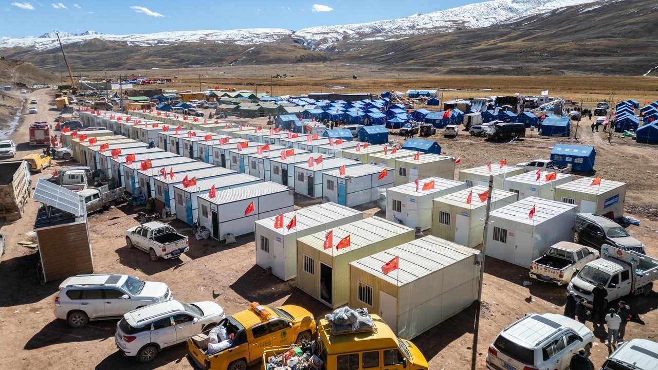Aerial photo, Oct. 25, 2026: rows of white prefabricated housing units with red flags and blue relief tents in Maru Township, Baqên County, after the earthquake, with snow-capped mountains behind
