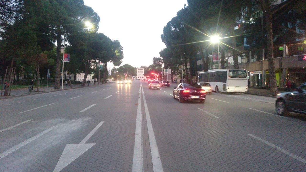 Stand-in, not a photo of this event: evening traffic on Dëshmorët e Kombit (Martyrs of the Nation) Boulevard in Tirana, Albania, with pine trees, lit street lamps, a white bus and cars in the lanes (October 2017)