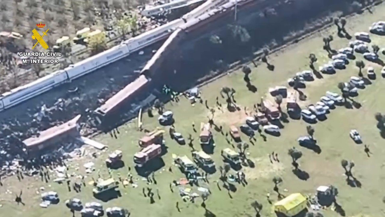 Still from an aerial video posted on X by @guardiacivil: derailed white and red high-speed train carriages lie across an embankment beside olive groves near Urda, Toledo, with debris on the grass and dozens of emergency vehicles and responders, too small to identify, parked in a field below; a Guardia Civil watermark sits at upper left