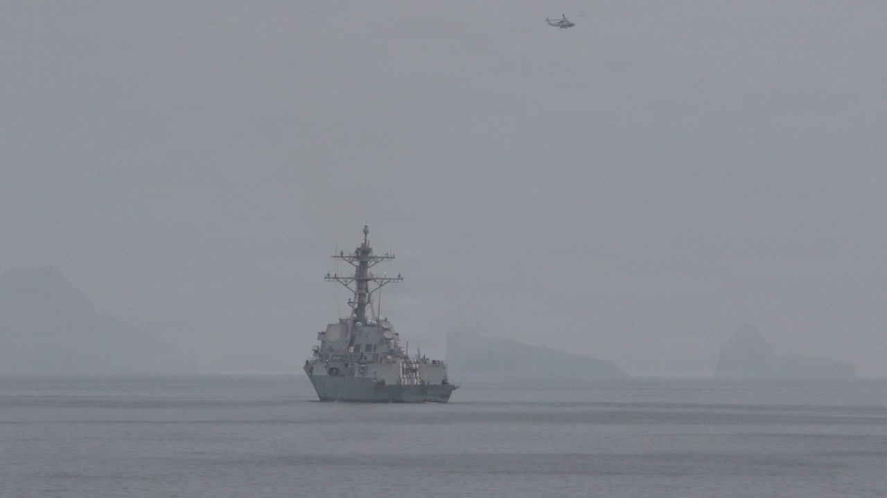 Stand-in, not a photo of this event: a gray U.S. Navy guided-missile destroyer on a calm, hazy sea in the Strait of Hormuz, with a helicopter overhead and faint rocky islands behind, in an April 11, 2026 U.S. Navy photo of mine-clearance operations