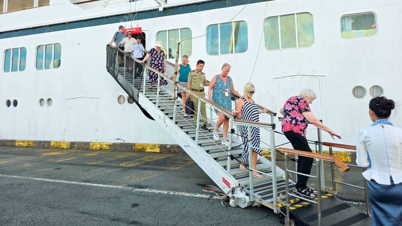 Photo posted on X by @jacobincambodia: passengers, several carrying bags, walk down a steep metal gangway from the white hull of a cruise ship onto a quayside, with a uniformed official among them and a woman in a pale uniform waiting at the foot of the stairs