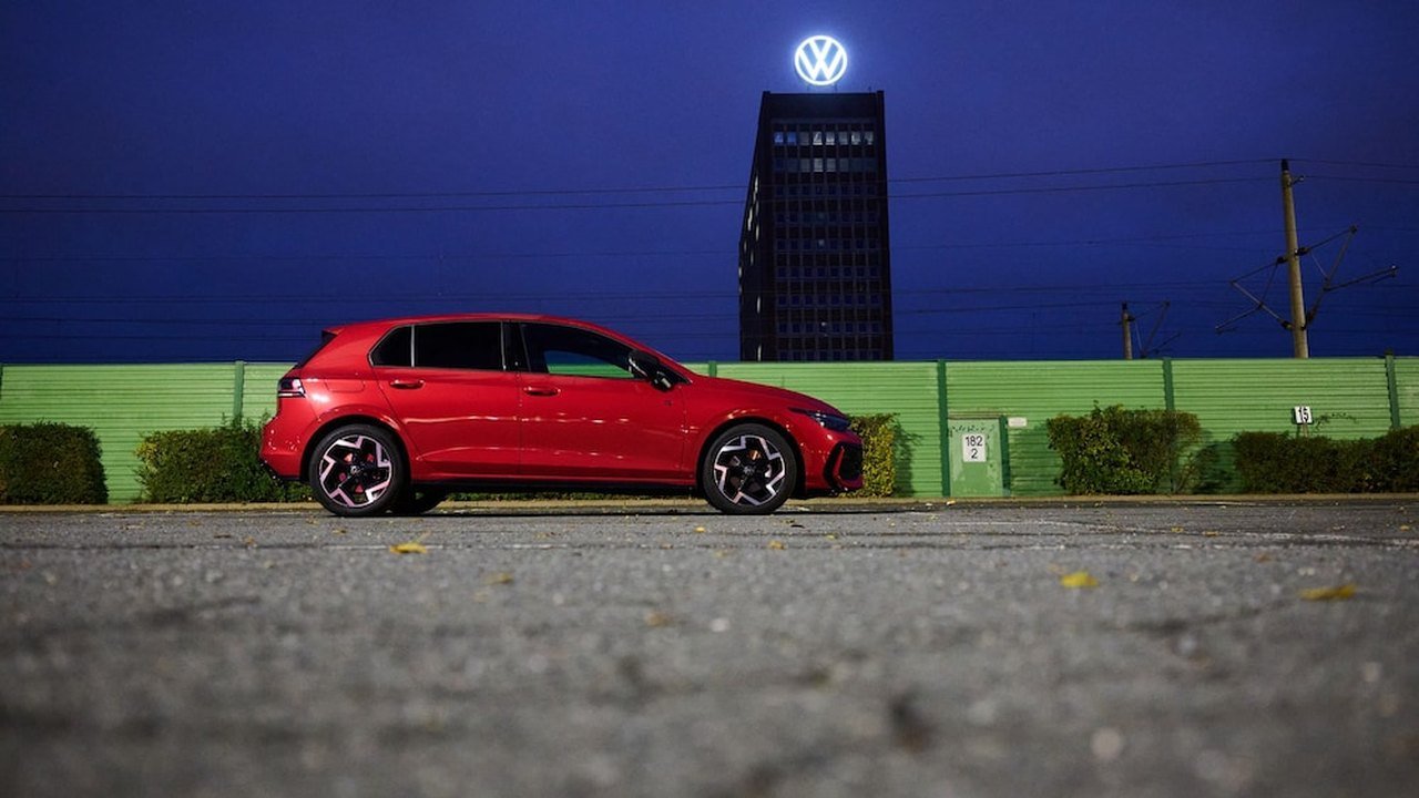A red Volkswagen Golf hatchback parked on asphalt in front of a green fence, with Volkswagen’s brand tower in Wolfsburg, topped by a lit VW logo, rising against a deep blue evening sky