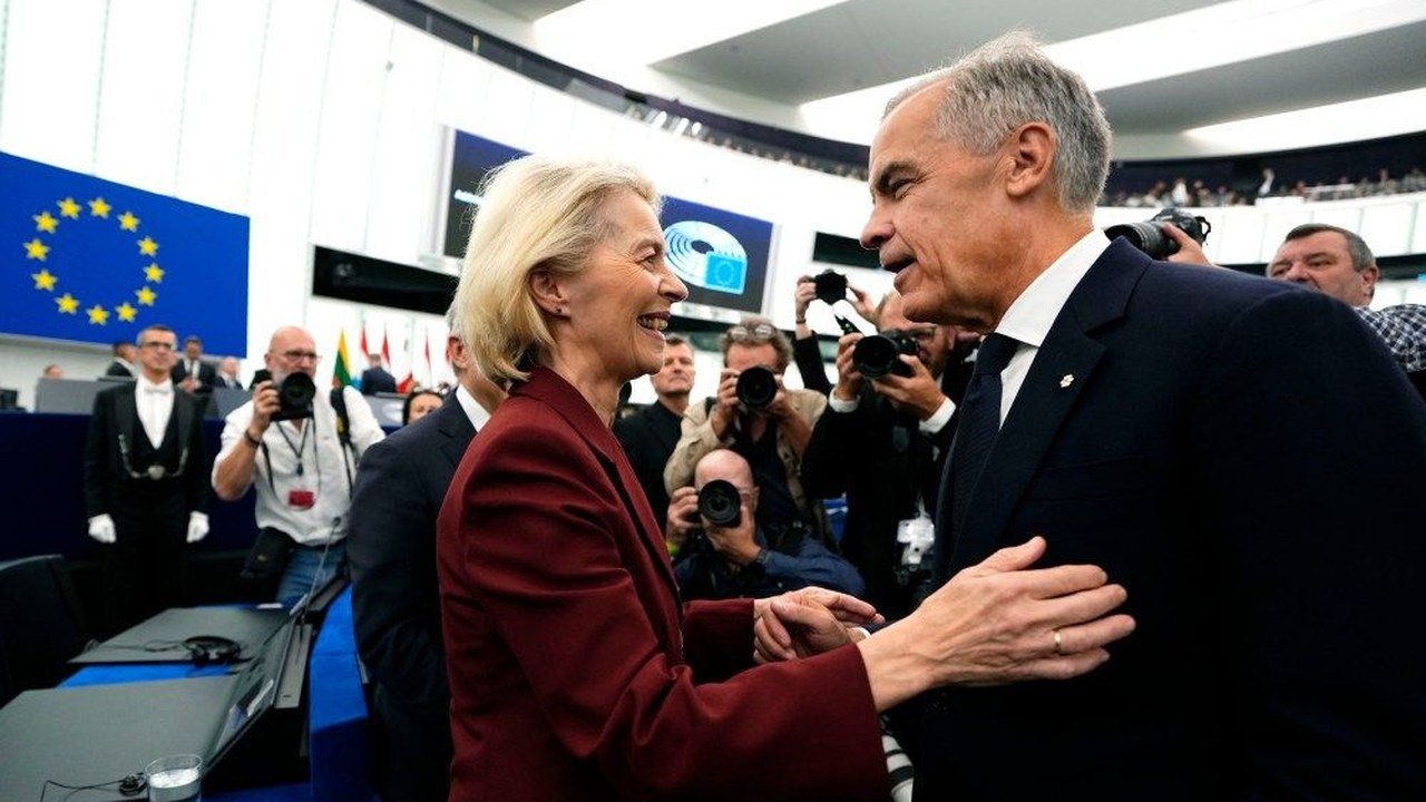 File image posted on X by @thebrandonsun: European Commission President Ursula von der Leyen, in a dark red jacket, smiles and clasps hands with Canadian Prime Minister Mark Carney inside a parliament chamber, with an EU flag on the left and photographers behind them; an earlier meeting, not the Montreal summit